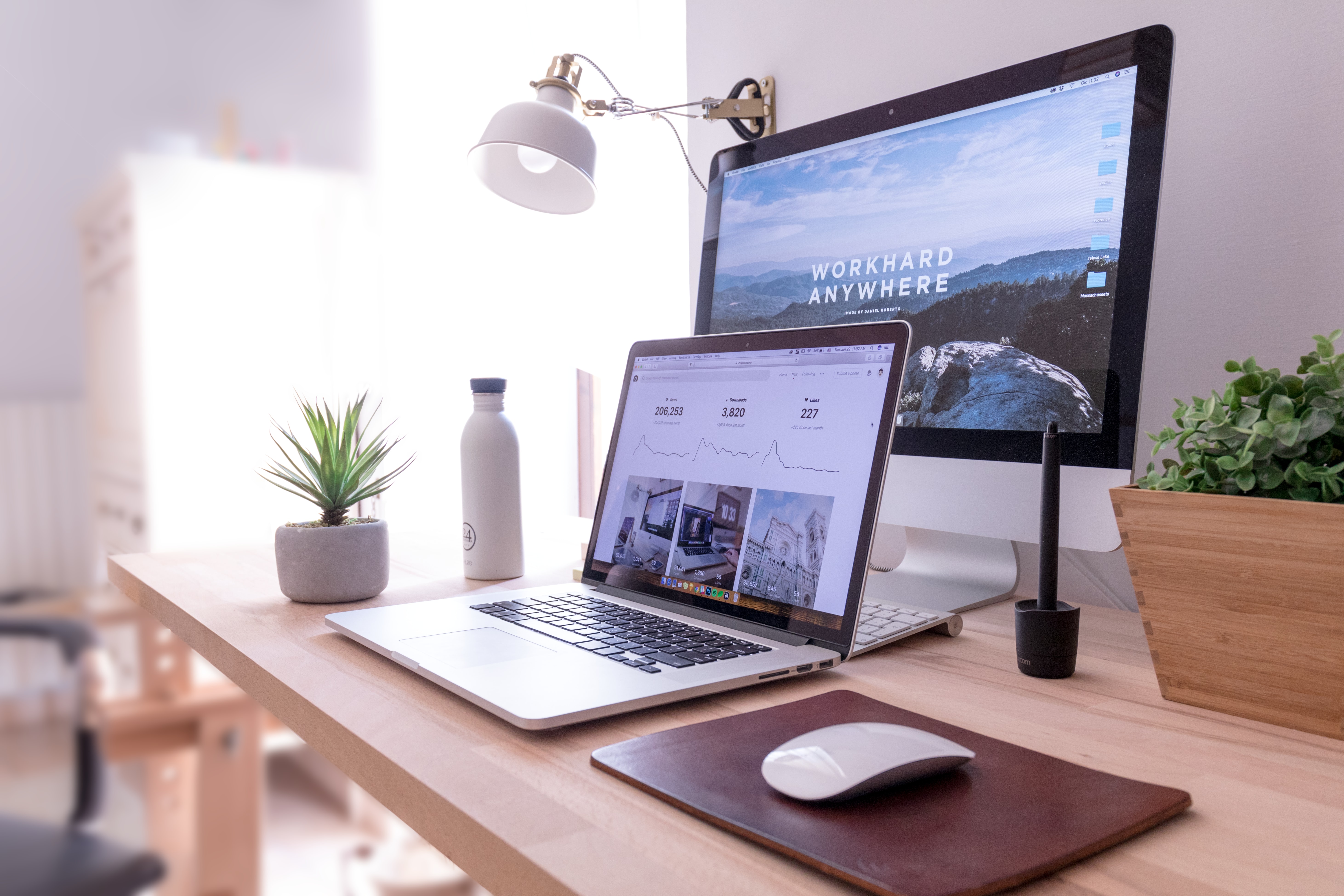 work from home desk with laptop, monitor and plant. Monitor displays message "work hard anywhere"